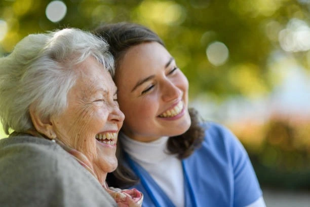 Une personne âgée sourie et passe un moment de convivialité avec une aide soignante souriante à ses côtés, tête contre tête.